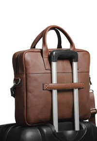 Sac en cuir marron avec deux poignées, texture lisse et fermeture éclair, reposant sur une valise noire avec une poignée télescopique.