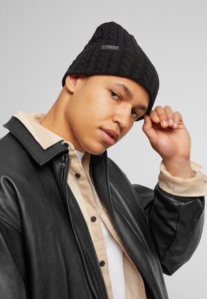 Mustal puuvillane beanie, millel on tekstuuriga muster ja silt, kus on kiri "CLASSICS." Kantud musta nahkjaki üle tan värvi nööpidega särgi.