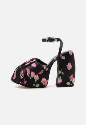 Sandale à talon haut plateforme noire avec bride à la cheville et motif floral de rose rose, avec un grand nœud sur la zone des orteils.