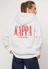 Mujer con coleta que lleva una sudadera blanca con capucha y texto rojo "KAPPA TORINO DAL 1967" en la espalda, y vaqueros azul oscuro, con las manos en los bolsillos.