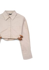 Camicia beige corta con colletto classico, maniche lunghe, tasca frontale e dettagli in finta pelle con fibbia in vita. Tessuto liscio.