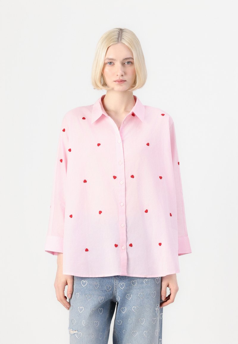 Ανοιχτό ροζ πουκάμισο με κόκκινο κεντημένο σχέδιο καρδιάς. Διαθέτει γιακά και μακριά, αναδιπλωμένα μανίκια. Χαλαρός σχεδιασμός με άνετη εφαρμογή.