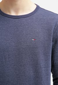 Detailní záběr na osobu oblečenou v tmavě modrém pleteném svetru s malým vyšitým logem červené, bílé a modré barvy na levé hrudi.