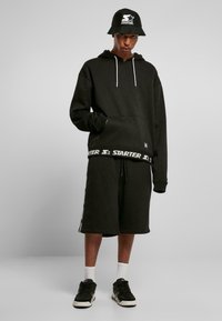 Hanorac negru cu glugă, dotat cu șnururi albe, purtat cu pantaloni scurți negri cu un pantalon cu logo alb la talie. Asociat cu adidași negri și o pălărie tip bucket.
