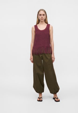 Jeune femme debout portant un haut sans manches bordeaux à franges, un pantalon ample vert olive avec des ourlets froncés, et des sandales noires à brides sur fond uni.