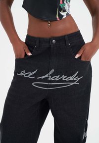 Svarta denimjeans med en framträdande grå "ed hardy" logotyp framtill, klassisk femfick-design och en figurnära midja.
