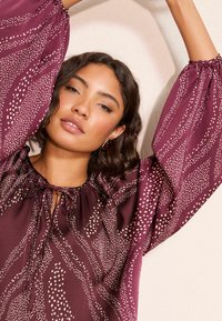 Burgunderfarbige Bluse mit gerafftem Ausschnitt, Bindeband und Puffärmeln. Sie verfügt über weiße Punktmuster und einen weichen, leichten Stoff.