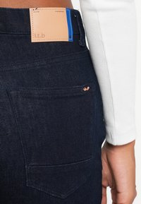 Jeans de mezclilla oscura con una etiqueta de cuero en la cintura, que presentan un bolsillo trasero y detalles de costura sutiles. Textura suave y diseño ajustado.