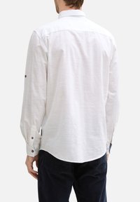 Chemise blanche à manches longues avec une texture douce, poignets à boutons et un ourlet arrondi. Le dos présente un col standard et des détails de couture subtils.