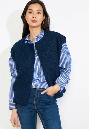 Frau in blau gestreiftem Hemd unter einer dunkelblauen ärmellosen Weste, kombiniert mit blauen Jeans, vor weißem Hintergrund stehend.