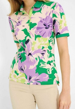 Nainen, jolla on vihreä, purppuranvärinen ja kerma kukkakuvioinen lyhythihainen poolopaita ja beiget housut, kuvattuna kaulasta lantioon.