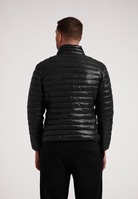 Giacca a vento nera con motivo a righe orizzontali, colletto alto e finitura lucida. Indossata sopra pantaloni scuri, mette in risalto una silhouette aderente.