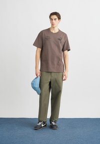 Καφέ κοντομάνικο μπλουζάκι με τυπωμένα γραφικά, συνδυασμένο με χακί παντελόνια. Κρατώντας ένα γαλάζιο καπέλο, φοράει μαύρα και λευκά αθλητικά παπούτσια.