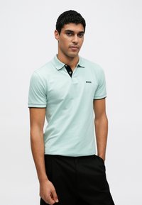 Világos menta zöld póló fekete gallérral és kétgombos gombolattal, bal oldali mellrészen kis fekete logóval. Rövid ujjú.