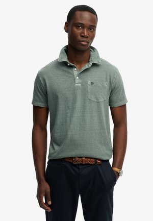 Mann trägt ein ausgewaschenes grünes Poloshirt mit kurzen Ärmeln und Tasche, geflochtener brauner Gürtel, dunkle Hose und Armbanduhr, steht mit einer Hand in der Tasche.