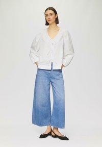Weißes Baumwollbluse mit V-Ausschnitt, Bindeband vorne und Puffärmeln, kombiniert mit hellblauen, weit geschnittenen Jeans. Schwarze flache Schuhe runden den Look ab.