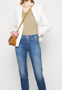 Woman wearing white blazer, beige top, blue jeans, gold necklace, and brown crossbody bag, standing against a white background.
