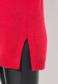 Czerwony sweter z dzianiny o luźnym kroju, z bocznymi rozcięciami i ribbowym wykończeniem. Tekstura wydaje się miękka i przewiewna, uzupełniająca gładką czarną dolną część.