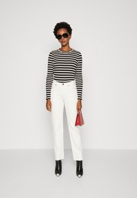 Maglietta a maniche lunghe a righe in bianco e nero, jeans bianchi a vita alta, stivaletti neri alla caviglia con punta bianca, che tiene una piccola borsa rossa.