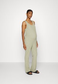 Macacão verde com texturização ribada, alças largas, cintura ajustada e calças flare. Combinado com sandálias pretas de sola plana. Design simples e casual.