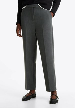 Grijze op maat gemaakte broek met pijpenlengte, gedragen met zwarte Mary Jane platte schoenen en een zwart topje met witte bies, handen nonchalant in de zakken.