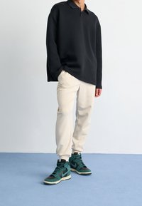 Černé polokošile s dlouhým rukávem, krémové zúžené tepláky a vysoké tenisky zelené a šedé barvy na modré podlaze před světlým pozadím.