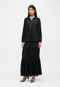 Μαύρη μακριμάνικη μπλούζα με σούρα στο ντεκολτέ και κορδόνι, συνδυασμένη με φούστα σε στρώσεις που ταιριάζει. Ομαλή υφή με διακριτικά μοτίβα.