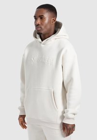 Sudadera oversized color crema con un bolsillo frontal, puños acanalados y logo en relieve. Tejido suave con una textura lisa, detalle de capucha con cordón.