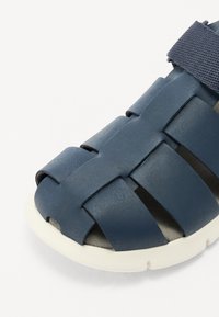 Sinivärvi kudumine sandaal tekstuuriga sünteetilisest pealmisest osast ja valgest kummist tallast. Omab reguleeritavat rihma kindlaks sobivuseks.