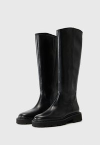 Botas de cuero negro hasta la rodilla con una textura suave, punteras redondas y una suela de goma gruesa. Presentan un diseño minimalista y líneas limpias.