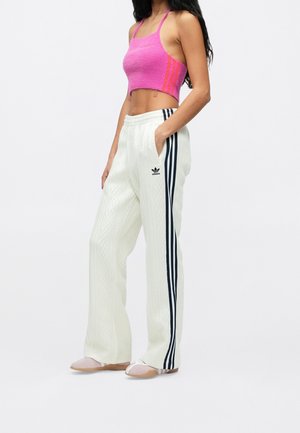 Vrouw draagt een roze cropped tanktop en witte Adidas wijde broek met zwarte strepen, staand tegen een effen achtergrond.