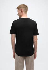 Schwarzes kurzärmliges T-Shirt mit rundem Ausschnitt, glatter Textur und etwas längerem Rückensaum, kombiniert mit hellen, locker sitzenden Hosen.