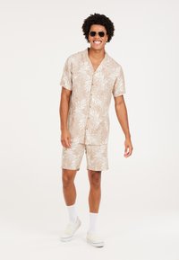 Camisa de manga corta y pantalones cortos de un tejido beige con un patrón de hojas blancas. Combinado con gafas de sol, calcetines blancos y zapatos tipo slip-on.