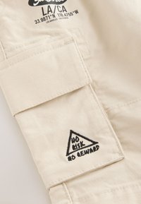 Beige fabric patch with a flap, featuring black embroidery of a triangular design and the text "NO RISK NO REWARD." Text above includes coordinates.