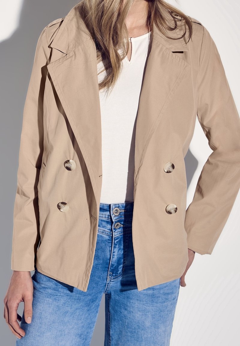 Femme portant un blazer beige à double boutonnage avec quatre boutons, un haut blanc et un jean bleu taille haute, debout avec une main sur la hanche.