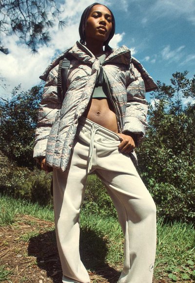 Pufferjacke met een gestructureerde metallic afwerking in grijstinten en roze, gecombineerd met lichtbeige sweatpants. Buitenomgeving met groenvoorzieningen.