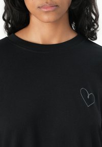 Sort bomulds t-shirt med rund hals og korte ærmer, der har et lille, lysegrå hjerte designet på venstre bryst. Blød tekstur.