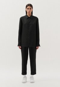 Camicia nera con bottoni e maniche lunghe, abbinata a pantaloni neri. Il modello indossa sneakers bianche e mostra una silhouette semplice e su misura.