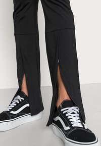 Pantalons de sport noirs avec zips latéraux, présentant une texture lisse. Associés à des baskets lacées noir et blanc affichant des accents contrastants.