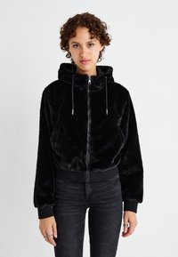 Zwarte nepbont zip-up hoodie met een aansluitende zoom, lange mouwen en verstelbare koordjes aan de capuchon. Gedragen met donkere jeans.