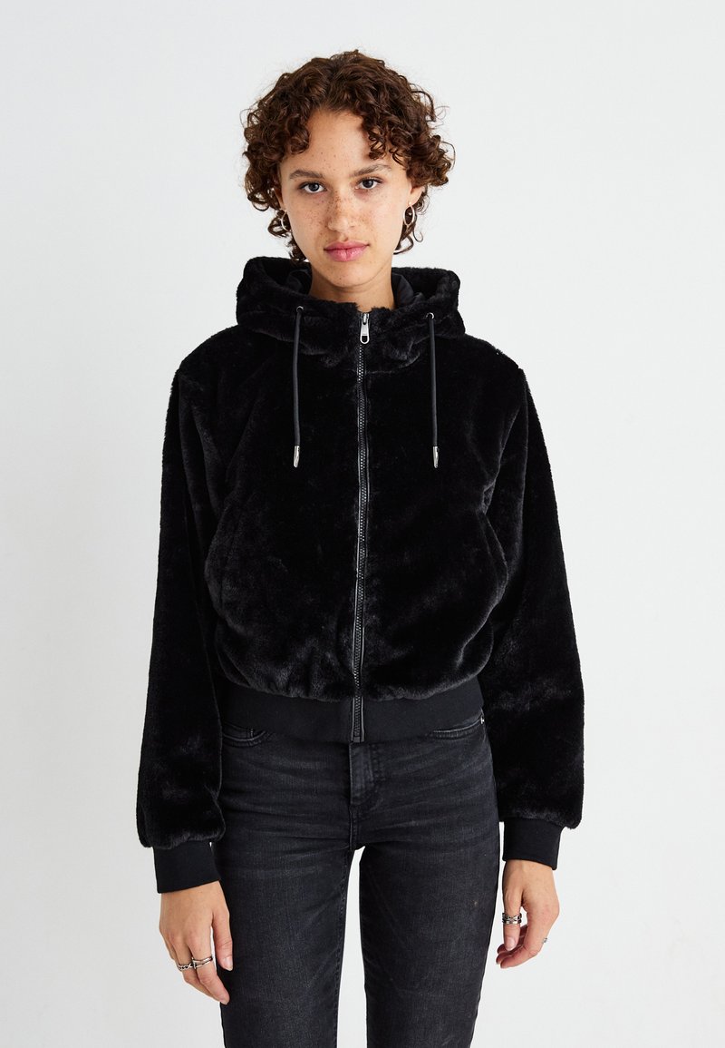 Zwarte nepbont zip-up hoodie met een aansluitende zoom, lange mouwen en verstelbare koordjes aan de capuchon. Gedragen met donkere jeans.