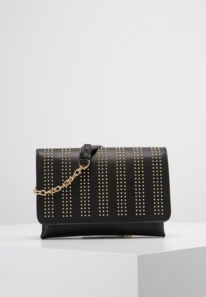 Schwarze Leder-Clutch mit senkrechten goldenen Nieten und kurzem goldenen Kettenriemen, liegend auf einem weißen Podest vor grauem Hintergrund.