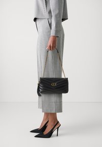 Borsa a spalla in pelle trapuntata nera con logo dorato e tracolla a catena, abbinata a un completo aderente a scacchi bianco e nero e tacchi a stiletto neri.