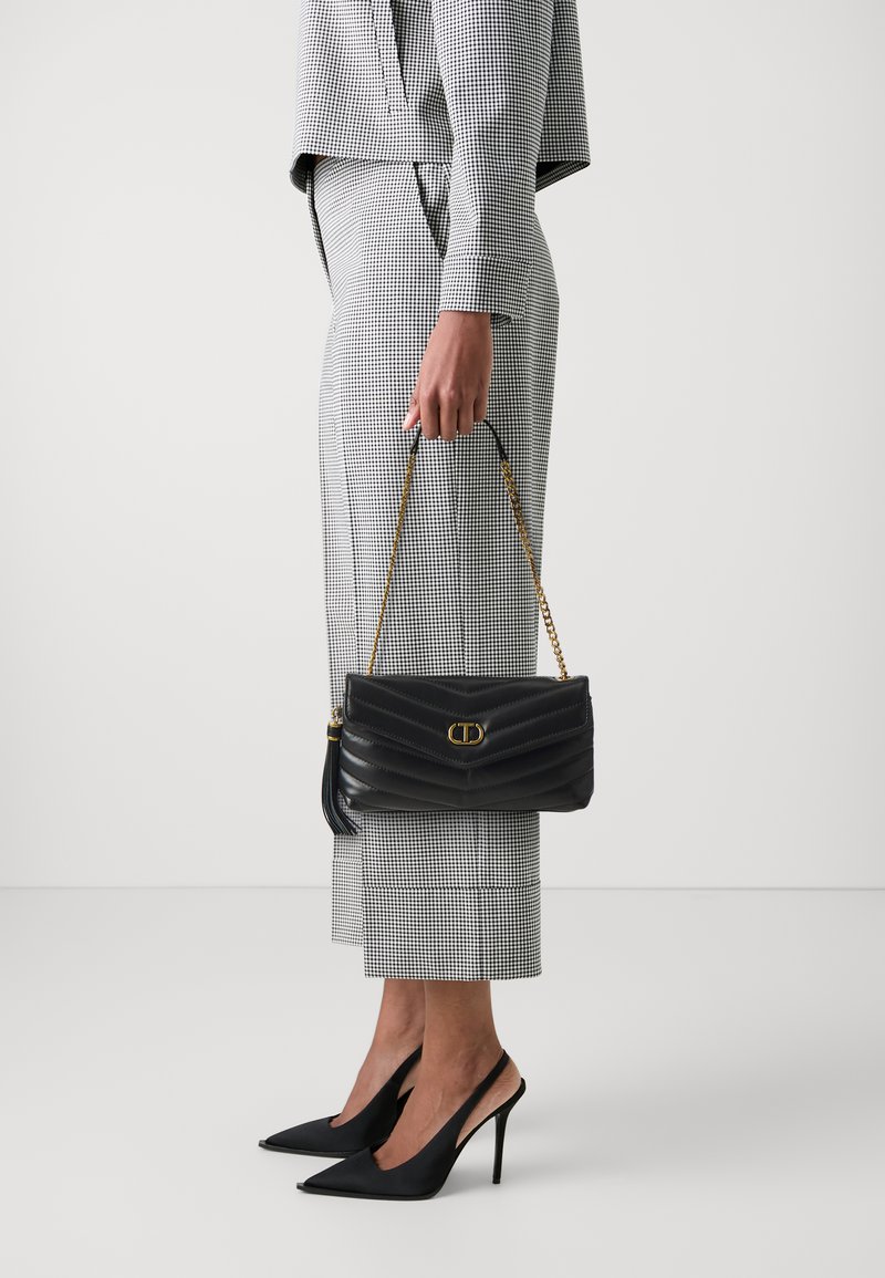 Borsa a spalla in pelle trapuntata nera con logo dorato e tracolla a catena, abbinata a un completo aderente a scacchi bianco e nero e tacchi a stiletto neri.