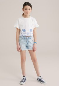 Camiseta de algodón blanca con bordado azul de palmeras y temática de playa, mangas con volantes, combinada con pantalones cortos de mezclilla claros y zapatillas navy.