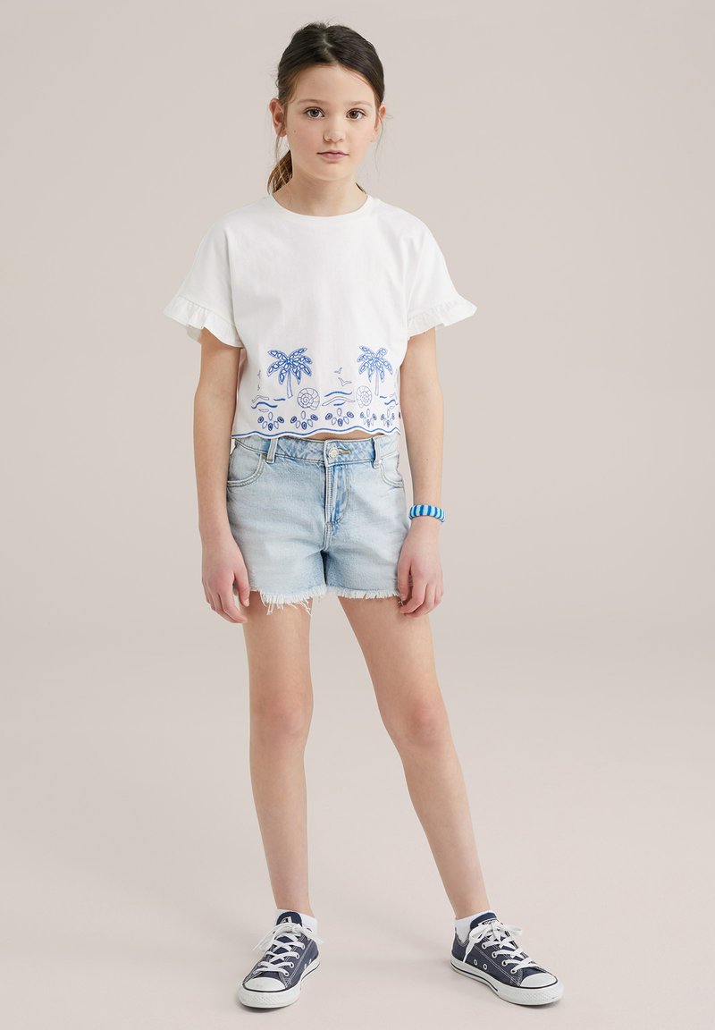 Camiseta de algodón blanca con bordado azul de palmeras y temática de playa, mangas con volantes, combinada con pantalones cortos de mezclilla claros y zapatillas navy.