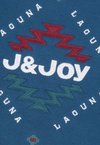 Blauwe stof met witte, rode en groene geborduurde tekst en zigzagpatronen, met de naam "J&Joy" centraal gepositioneerd.