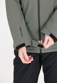 Grüne wasserdichte Jacke mit verstellbaren Klettverschlussärmeln. Sie bietet ein elegantes Design mit einem Reißverschluss und der „W-PRO 10000“-Markierung am Ärmel.