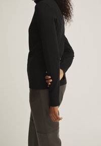 Svart stickad tröja med hög krage och långa ärmar, i kombination med bruna byxor i konstläder. Slät textur, figursydd design.