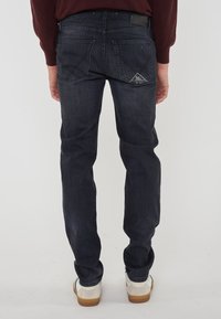 Uomo che indossa jeans blu scuri, un maglione bordeaux e sneaker bianche, in piedi con la schiena rivolta verso la fotocamera su uno sfondo bianco uniforme.